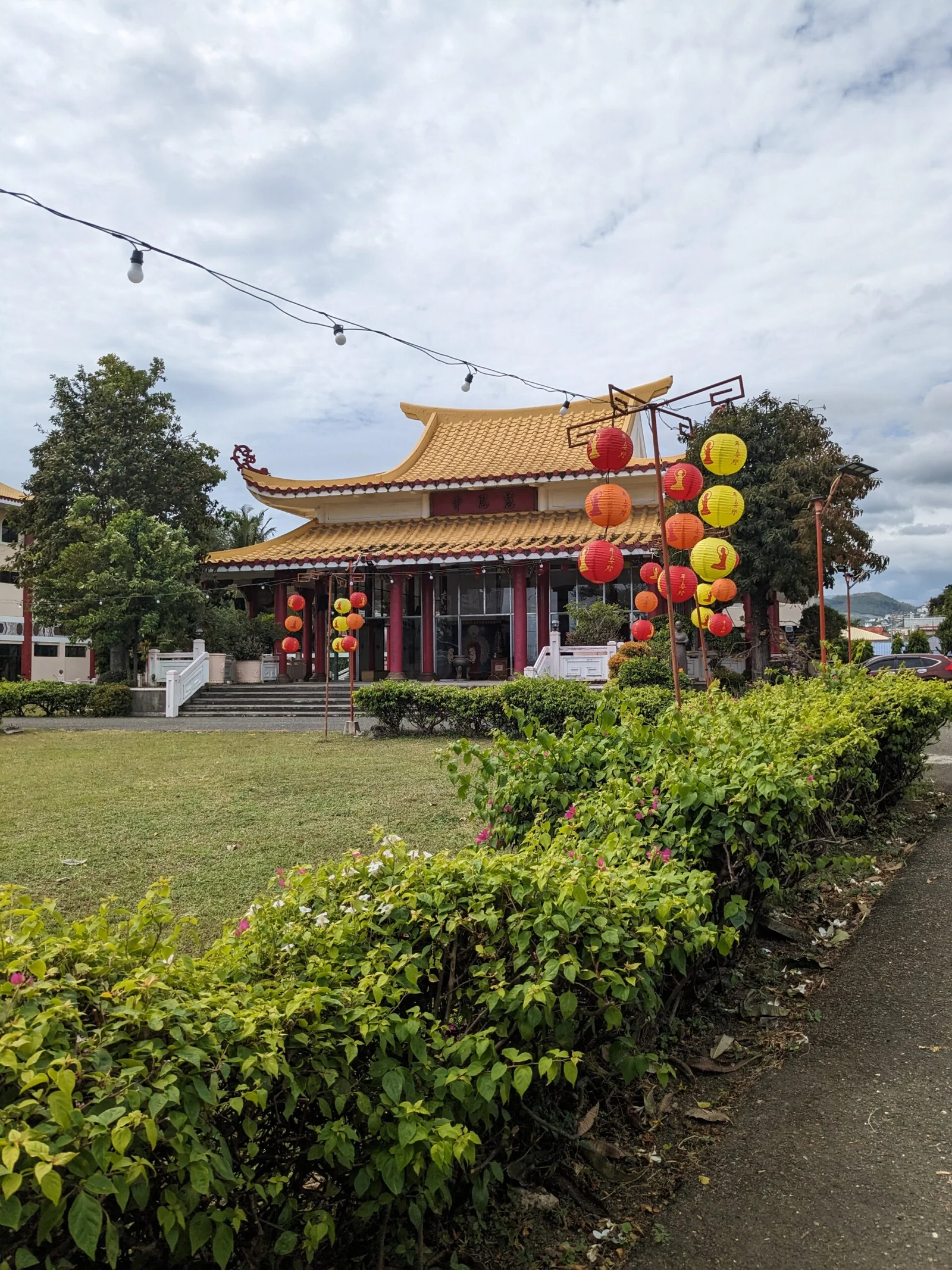 A traditional Chinese temple with a yellow roof, red columns, and hanging red and yellow lanterns, surrounded by greenery and a cloudy sky.